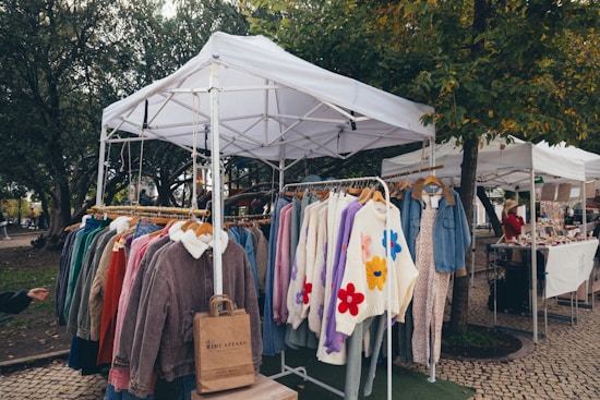 A market stall featuring a collection of colorful winter clothing items displayed on hangers under a white canopy. The garments include jackets and sweaters, some with floral patterns. The scene is set in an outdoor environment surrounded by trees and cobblestone paths, with another stall and a person visible in the background.