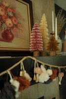 A cozy and artistic interior setting featuring a painting of a flower bouquet in a vase on the wall. Below the painting, there are decorative miniature Christmas trees in pink, golden, and yellow colors, adorned with white tips to resemble snow. Next to the trees are elegant white candles in holders. A charming garland with soft fabric figures and bells is draped on the mantle, adding a festive touch to the decor.