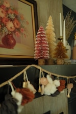 A cozy and artistic interior setting featuring a painting of a flower bouquet in a vase on the wall. Below the painting, there are decorative miniature Christmas trees in pink, golden, and yellow colors, adorned with white tips to resemble snow. Next to the trees are elegant white candles in holders. A charming garland with soft fabric figures and bells is draped on the mantle, adding a festive touch to the decor.