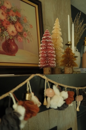 A cozy and artistic interior setting featuring a painting of a flower bouquet in a vase on the wall. Below the painting, there are decorative miniature Christmas trees in pink, golden, and yellow colors, adorned with white tips to resemble snow. Next to the trees are elegant white candles in holders. A charming garland with soft fabric figures and bells is draped on the mantle, adding a festive touch to the decor.