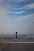 A person stands alone on a concrete platform overlooking the sea, with a cloudy sky above and a distant horizon. The person is wearing a dark jacket and jeans while carrying a backpack. The image conveys a sense of solitude and contemplation.