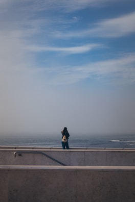 A person stands alone on a concrete platform overlooking the sea, with a cloudy sky above and a distant horizon. The person is wearing a dark jacket and jeans while carrying a backpack. The image conveys a sense of solitude and contemplation.