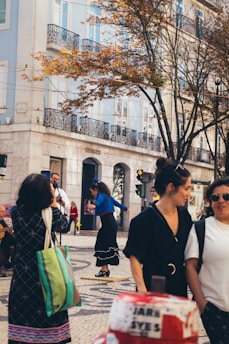 A street scene in a city with people walking and a woman dancing on a wooden platform. The background features a pastel-colored building with balconies and a tree with autumn leaves. Pedestrians are captured mid-motion, and a street musician plays a guitar.