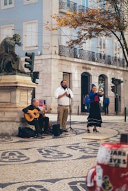 Three street performers are seen in a vibrant urban setting. One musician is playing a guitar while seated beside a large statue. A man stands at a microphone and appears to be singing. A woman, dressed in traditional clothing and wearing a blue headscarf, is passionately performing, possibly dancing or singing. The backdrop includes an old stone building with balconies and large windows, with autumn foliage on trees providing a contrast of colors.