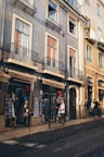 A picturesque European street with elegant buildings featuring intricate wrought-iron balconies and large windows. The shops on the ground floor display various wares, including hats and souvenirs. Several people are strolling past the shops, and the ambiance is relaxed and leisurely.