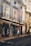 A picturesque European street with elegant buildings featuring intricate wrought-iron balconies and large windows. The shops on the ground floor display various wares, including hats and souvenirs. Several people are strolling past the shops, and the ambiance is relaxed and leisurely.