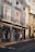 A picturesque European street with elegant buildings featuring intricate wrought-iron balconies and large windows. The shops on the ground floor display various wares, including hats and souvenirs. Several people are strolling past the shops, and the ambiance is relaxed and leisurely.