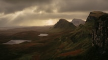 The image depicts a dramatic and expansive landscape with rolling hills, rugged cliffs, and scattered patches of greenery. A moody sky filled with dense, dark clouds allows rays of soft sunlight to break through, illuminating parts of the terrain. Small lakes are nestled between the hills, reflecting the subdued light.
