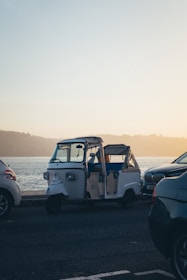 A three-wheeled vehicle, possibly a tuk-tuk or auto-rickshaw, is parked between two cars along a waterfront. The scene is set during what appears to be early morning or late afternoon, with a subtle glow from the sun reflecting off the water and casting a warm hue across the image.