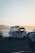 A three-wheeled vehicle, possibly a tuk-tuk or auto-rickshaw, is parked between two cars along a waterfront. The scene is set during what appears to be early morning or late afternoon, with a subtle glow from the sun reflecting off the water and casting a warm hue across the image.