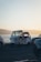 A three-wheeled vehicle, possibly a tuk-tuk or auto-rickshaw, is parked between two cars along a waterfront. The scene is set during what appears to be early morning or late afternoon, with a subtle glow from the sun reflecting off the water and casting a warm hue across the image.