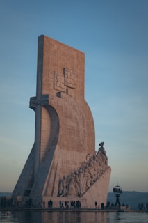 A large stone monument featuring a group of people sculpted along its side, appearing to be carved into the structure. The monument is positioned near a body of water with reflections visible on the water's surface. People are walking and observing around the base of the monument.