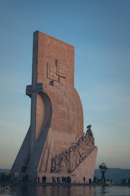 A large stone monument featuring a group of people sculpted along its side, appearing to be carved into the structure. The monument is positioned near a body of water with reflections visible on the water's surface. People are walking and observing around the base of the monument.