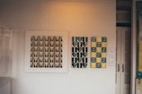 A room showcases three pieces of modern art on a neutral-toned wall. The leftmost artwork features a pattern of black and white geometric triangles. The central piece displays a pattern of teal and gray hearts against a striped background. On the right, a composition of yellow and blue abstract shapes with black stripes is presented.