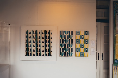 A room showcases three pieces of modern art on a neutral-toned wall. The leftmost artwork features a pattern of black and white geometric triangles. The central piece displays a pattern of teal and gray hearts against a striped background. On the right, a composition of yellow and blue abstract shapes with black stripes is presented.