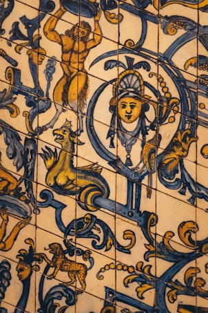 A detailed and colorful tile artwork featuring various mythological and decorative elements. At the center, there is a face wearing an ornate headdress, surrounded by intricate blue and yellow motifs. A lion-like mythical creature with exaggerated features is depicted alongside a lively scene involving a muscular figure holding an object. Additional scrolls and ornamental flourishes fill the design.