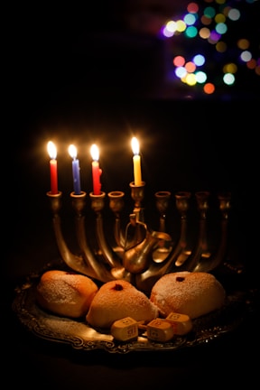 A menorah with five lit candles is set on a table, accompanied by sufganiyot, traditional doughnuts, dusted with sugar, and dreidels. In the background, colorful bokeh lights create a festive atmosphere.