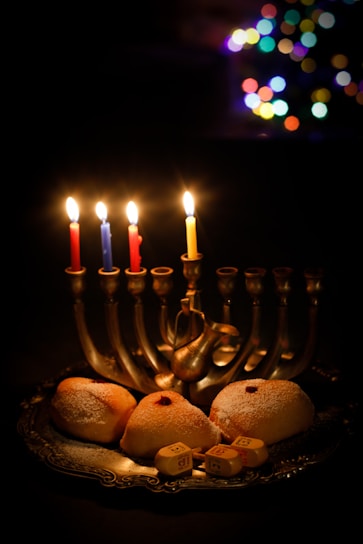 A menorah with five lit candles is set on a table, accompanied by sufganiyot, traditional doughnuts, dusted with sugar, and dreidels. In the background, colorful bokeh lights create a festive atmosphere.