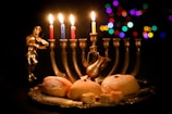 A lit menorah with five candles, surrounded by a small figurine playing a violin, a silver decorative pitcher, and a plate of three powdered doughnuts. The background is dark with colorful out-of-focus lights creating a festive atmosphere.