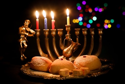 A lit menorah with five candles, surrounded by a small figurine playing a violin, a silver decorative pitcher, and a plate of three powdered doughnuts. The background is dark with colorful out-of-focus lights creating a festive atmosphere.