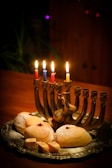 A silver menorah with lit candles in different colors sits on a wooden table, surrounded by freshly baked pastries sprinkled with powdered sugar, and small wooden spinning tops, called dreidels.
