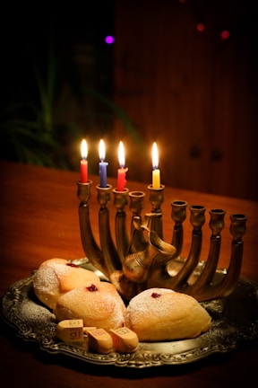 A silver menorah with lit candles in different colors sits on a wooden table, surrounded by freshly baked pastries sprinkled with powdered sugar, and small wooden spinning tops, called dreidels.