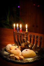 A silver menorah with lit candles in different colors sits on a wooden table, surrounded by freshly baked pastries sprinkled with powdered sugar, and small wooden spinning tops, called dreidels.