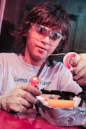 A person wearing protective goggles and gloves is holding a soldering iron and soldering wire. They are working on an electronic project, which is placed in a container covered with duct tape.
