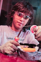 A person wearing protective goggles and gloves is holding a soldering iron and soldering wire. They are working on an electronic project, which is placed in a container covered with duct tape.