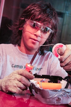 A person wearing protective goggles and gloves is holding a soldering iron and soldering wire. They are working on an electronic project, which is placed in a container covered with duct tape.