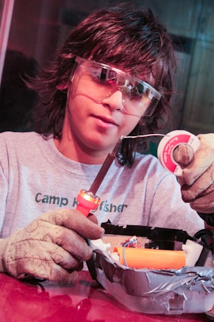A person wearing protective goggles and gloves is holding a soldering iron and soldering wire. They are working on an electronic project, which is placed in a container covered with duct tape.