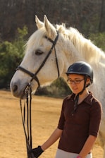 A young person wearing a helmet stands beside a large white horse, holding its reins. The horse appears calm and relaxed, with its fur visibly soft and well-groomed. Behind them, there is a natural setting with trees and the ground is covered with dry grass.