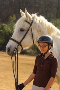 A young person wearing a helmet stands beside a large white horse, holding its reins. The horse appears calm and relaxed, with its fur visibly soft and well-groomed. Behind them, there is a natural setting with trees and the ground is covered with dry grass.