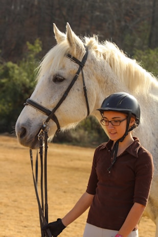 A young person wearing a helmet stands beside a large white horse, holding its reins. The horse appears calm and relaxed, with its fur visibly soft and well-groomed. Behind them, there is a natural setting with trees and the ground is covered with dry grass.
