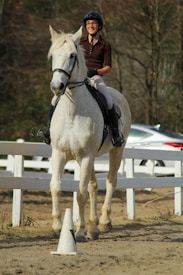 A person wearing a helmet and riding a white horse in a fenced outdoor riding area. The rider appears to be enjoying the ride, with a slight smile. The background includes a white fence and some blurry trees, suggesting an outdoor setting.