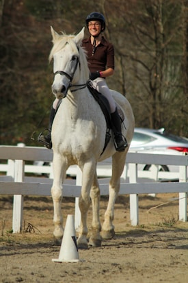 A person wearing a helmet and riding a white horse in a fenced outdoor riding area. The rider appears to be enjoying the ride, with a slight smile. The background includes a white fence and some blurry trees, suggesting an outdoor setting.