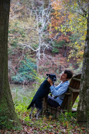 A person sits on a wooden chair in a forested area, affectionately playing with a black and white dog. The background features a serene pond surrounded by trees with autumn foliage, displaying a mix of green, orange, and brown leaves. The scene is peaceful and natural, with soft lighting filtering through the trees.