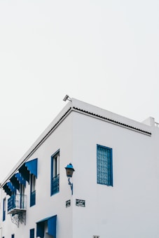 A white building with distinct blue accents, including window frames, shutters, and a small balcony. The architectural style appears Mediterranean, with clean lines and decorative metalwork on the windows. A blue street lamp is attached to the corner of the building, and two street signs are visible.