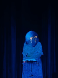 A mysterious figure with a blurred face, appearing like a globe, is standing behind a lectern with an open book. The scene is enveloped in a deep blue light, creating an enigmatic and surreal atmosphere.