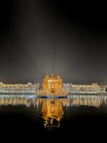 A beautifully illuminated golden temple is centrally positioned, surrounded by a large water body reflecting its glowing structure. The night sky serves as a backdrop, enhancing the temple's radiant appearance. It is accompanied by a long row of similarly lit buildings and decorative lights in the background.