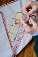 Hands are performing embroidery work on a piece of fabric that has a stitched border and colorful geometric patterns. A spool of thread is visible in the center of the fabric.