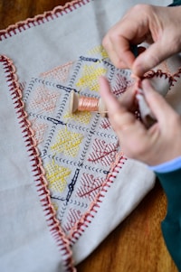 Hands are performing embroidery work on a piece of fabric that has a stitched border and colorful geometric patterns. A spool of thread is visible in the center of the fabric.
