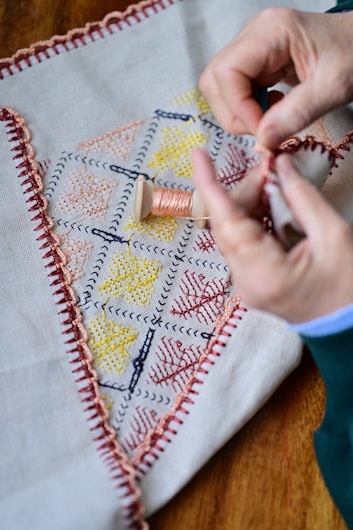 Hands are performing embroidery work on a piece of fabric that has a stitched border and colorful geometric patterns. A spool of thread is visible in the center of the fabric.