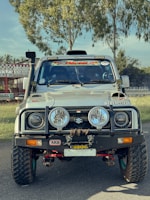A rugged, modified white Suzuki vehicle is parked on a road. It features large off-road tires, additional front bumper accessories, and various decals on the windshield and hood. The background consists of green trees and a racing track.