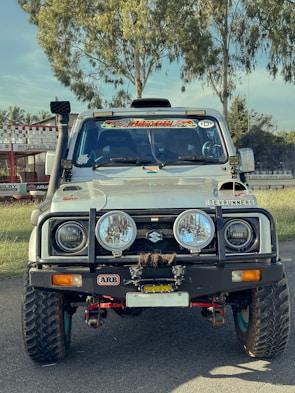 A rugged, modified white Suzuki vehicle is parked on a road. It features large off-road tires, additional front bumper accessories, and various decals on the windshield and hood. The background consists of green trees and a racing track.