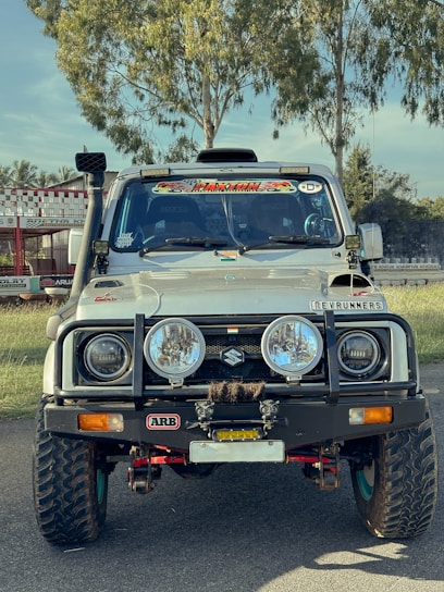 A rugged, modified white Suzuki vehicle is parked on a road. It features large off-road tires, additional front bumper accessories, and various decals on the windshield and hood. The background consists of green trees and a racing track.