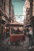 A street vendor with a red cart is selling chestnuts in an urban alleyway lined with tall buildings. A person stands nearby, engaged with the vendor. The scene is bustling with a few people visible in the background, captured in a warm, nostalgic atmosphere.