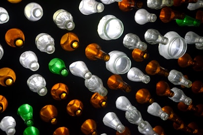 A collection of variously sized and shaped glass bottles arranged on a dark surface. The bottles are predominantly transparent, with some green and amber ones interspersed. The bottles are illuminated, creating reflections and highlighting their textures.