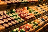 Shelves are filled with an array of colorful, packaged cheese wheels. The cheese wheels are neatly arranged and displayed under warm lighting, enhancing their vibrant colors like green, red, orange, and purple.