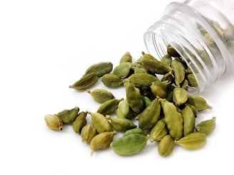 A clear plastic container is tipped over, spilling out numerous green cardamom pods onto a white surface. The pods are oblong and have a slightly ribbed texture.